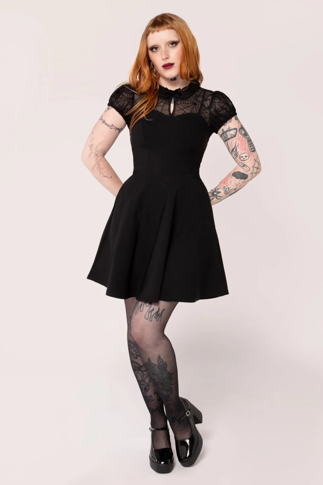 A model wearing a black mini dress with mesh fabric on the chest and sleeves, and velvet flocked spider webs on the chest and waistline. The dress has a sweetheart neckline and a fitted bodice with a skirt that flares out at the waistline.