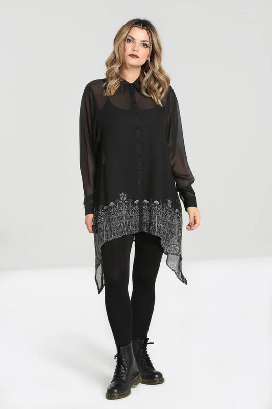 A woman wearing a black chiffon tunic with a print of ornate gate along the hem, featuring a contrasting black pointed collar and cuffs, and black buttons. She is also wearing black boots and leggings.